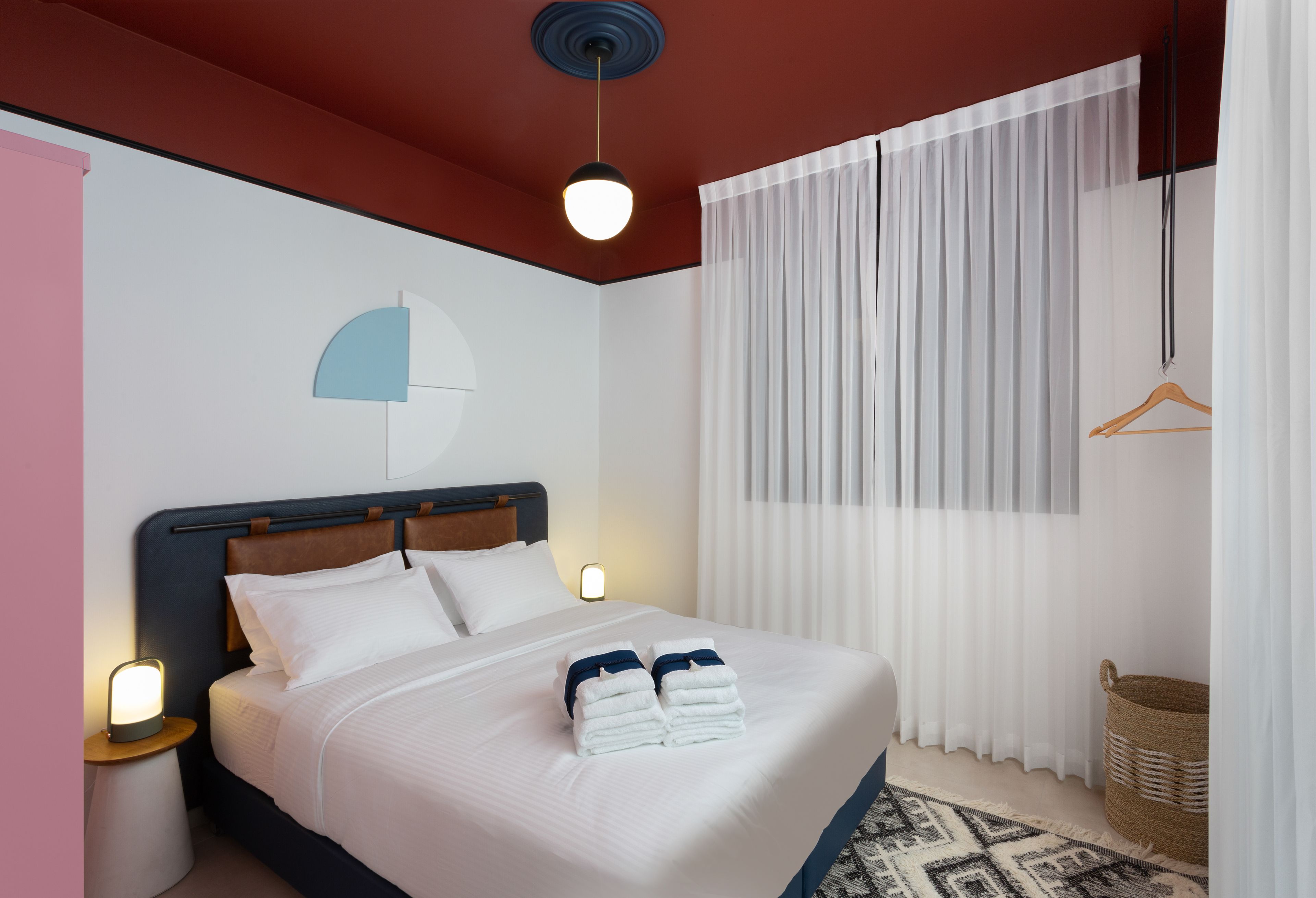 A neatly made bed with white linens and folded towels, a bedside lamp, and a wall-mounted light fixture.