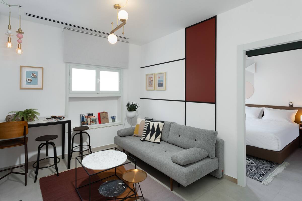 Une pièce moderne et bien éclairée, avec un canapé gris, une petite table ronde, un bureau avec deux tabourets, un lit doté d’une tête de lit blanche et une grande fenêtre.