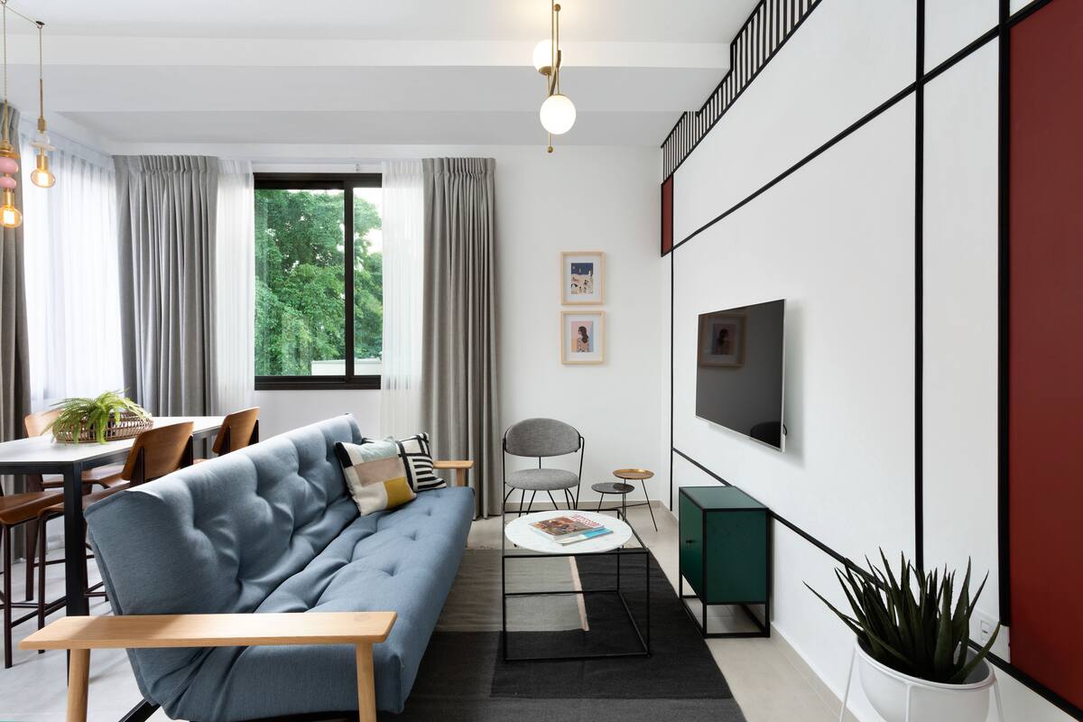 Un salon moderne comprenant un canapé, une table à manger et des chaises, un téléviseur fixé au mur et une grande fenêtre avec des rideaux.