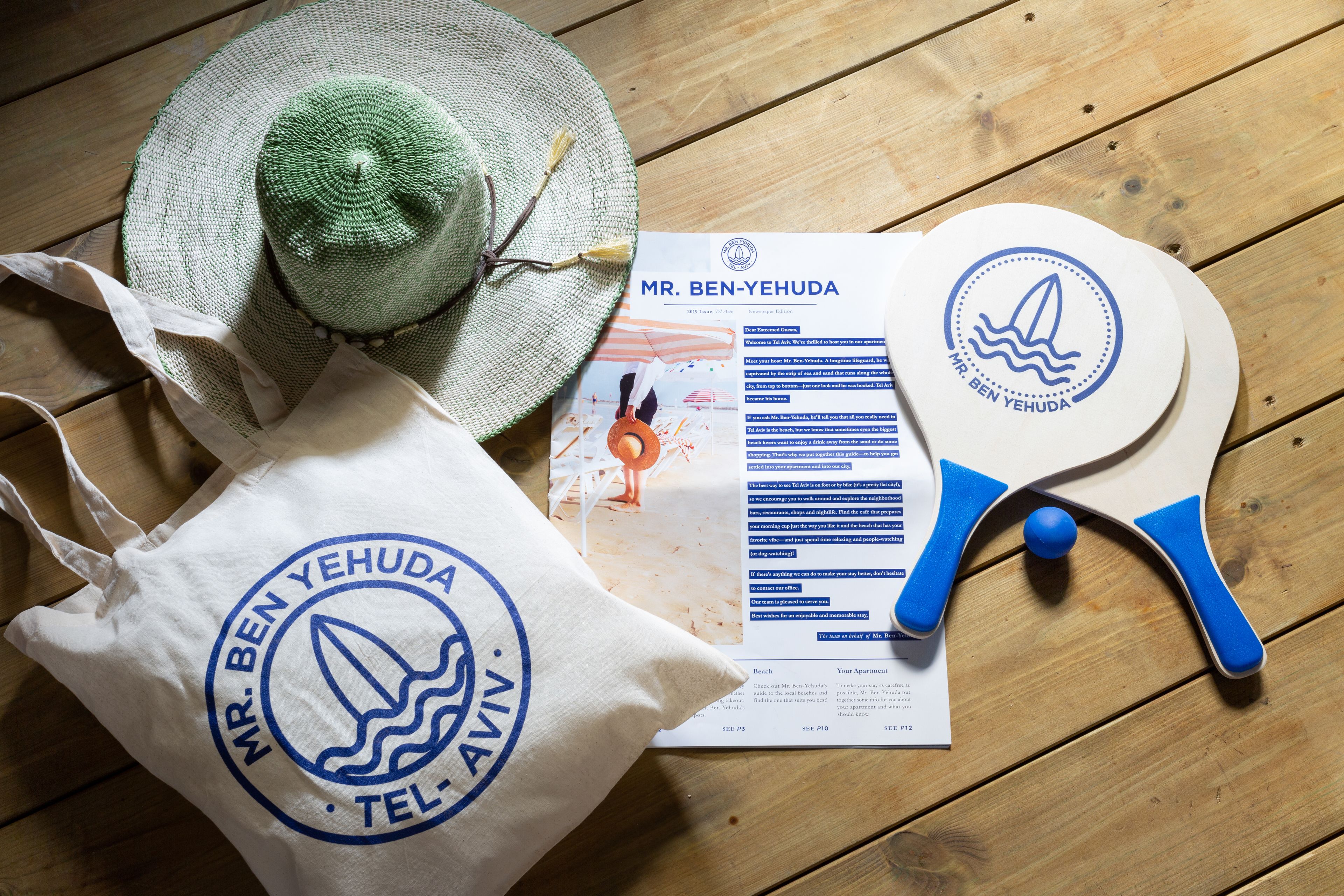 A wooden table with a green hat, a white tote bag with a logo, and a paddleboard.