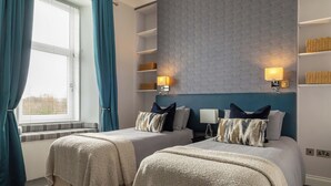 Habitación de hotel con dos camas, una ventana con cortinas, una pared con estantes y dos lámparas fijadas a la pared.