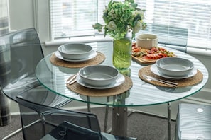 Une table en verre sur laquelle reposent quatre bols blancs, un vase avec des fleurs vertes et une planche en bois avec de la nourriture.