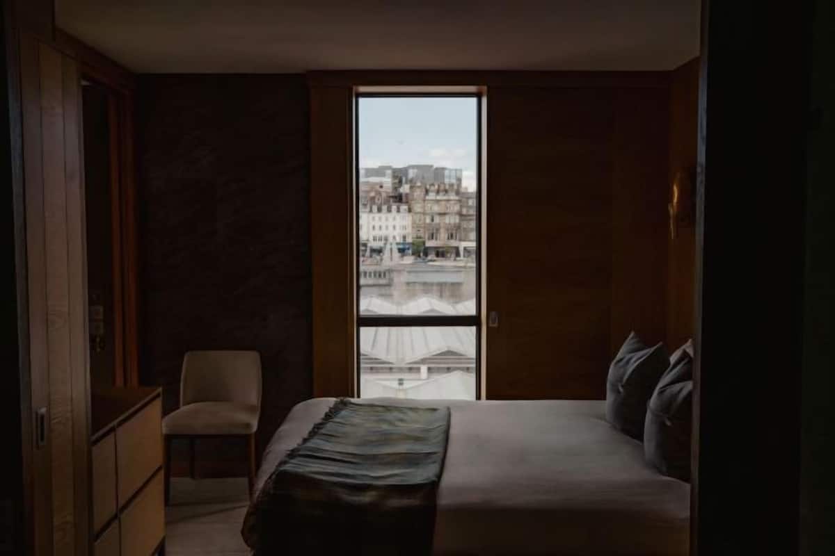 A bedroom with a bed, a chair, a wardrobe, and a large window with a city view.