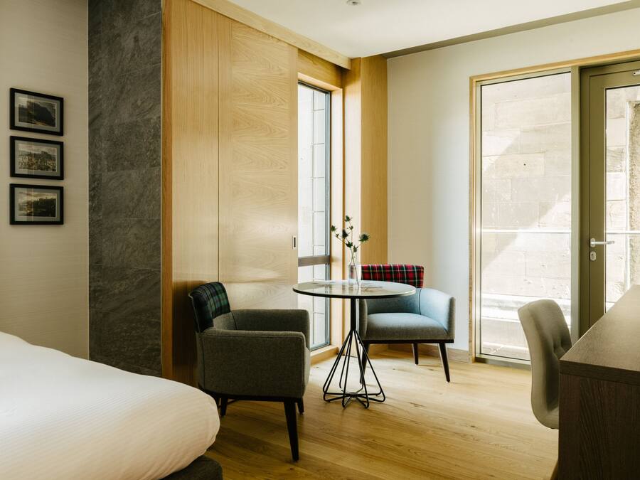 A hotel room with a bed, two armchairs, a small table, and a large window.