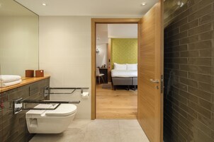 Een moderne badkamer met toilet, wastafel en handdoekhouder. Een houten deur leidt naar een slaapkamer met een bed en nachtkastje.