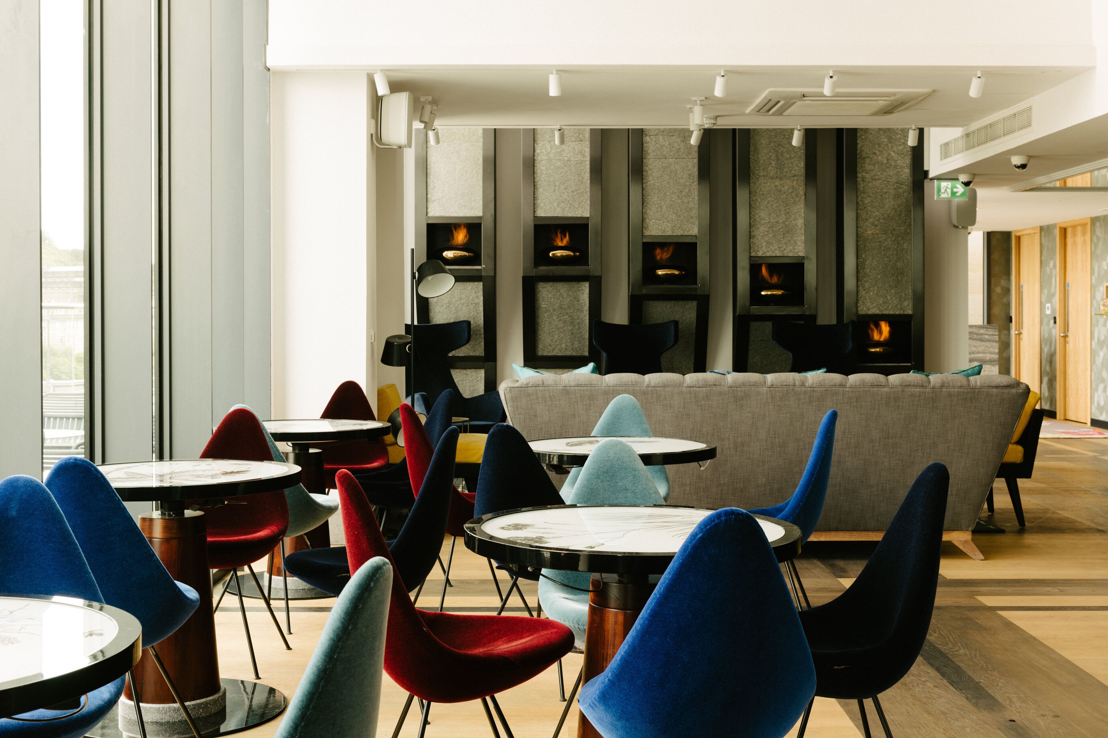 A modern interior with round tables and colorful chairs, a sofa, and a fireplace with flames.