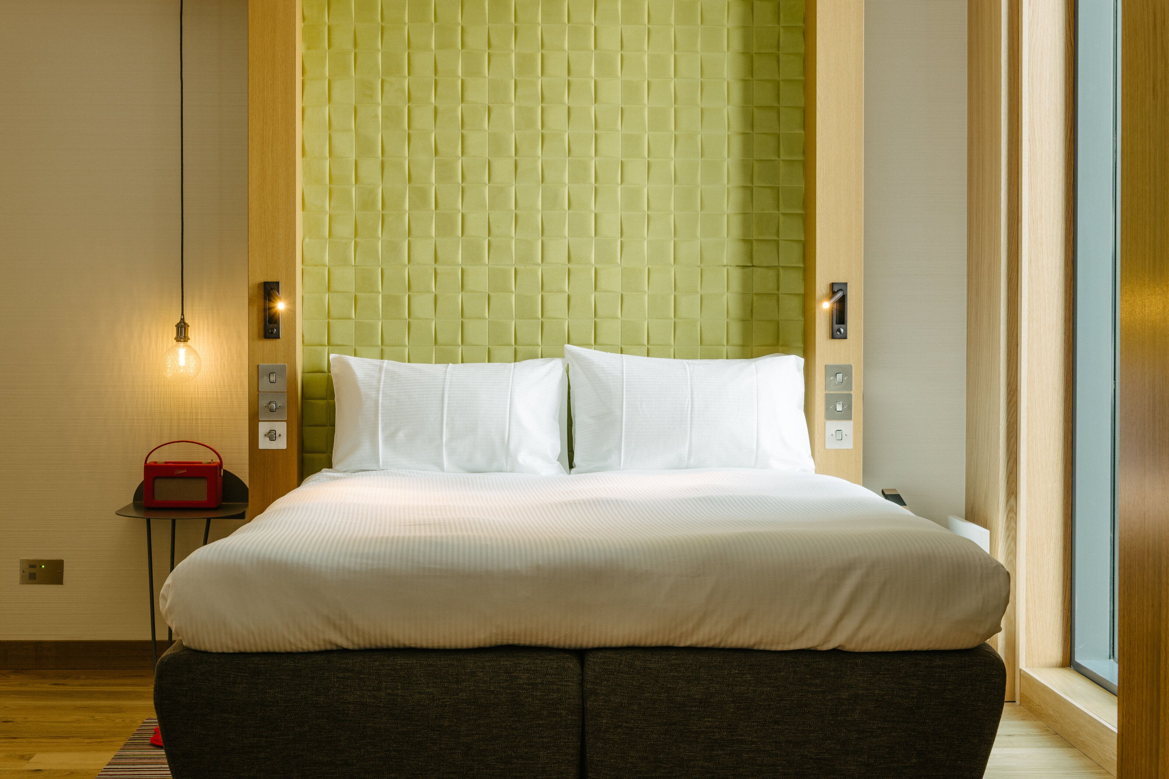 Una cama bien hecha con ropa blanca, una pared verde con textura y una mesita de noche con una radio roja al lado.