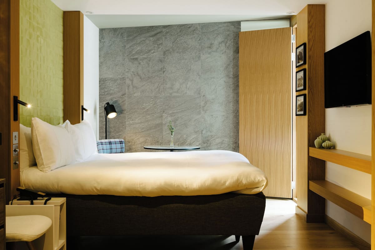 A modern bedroom with a large bed, bedside tables, a small round table, a wall-mounted TV, and a wooden shelf.