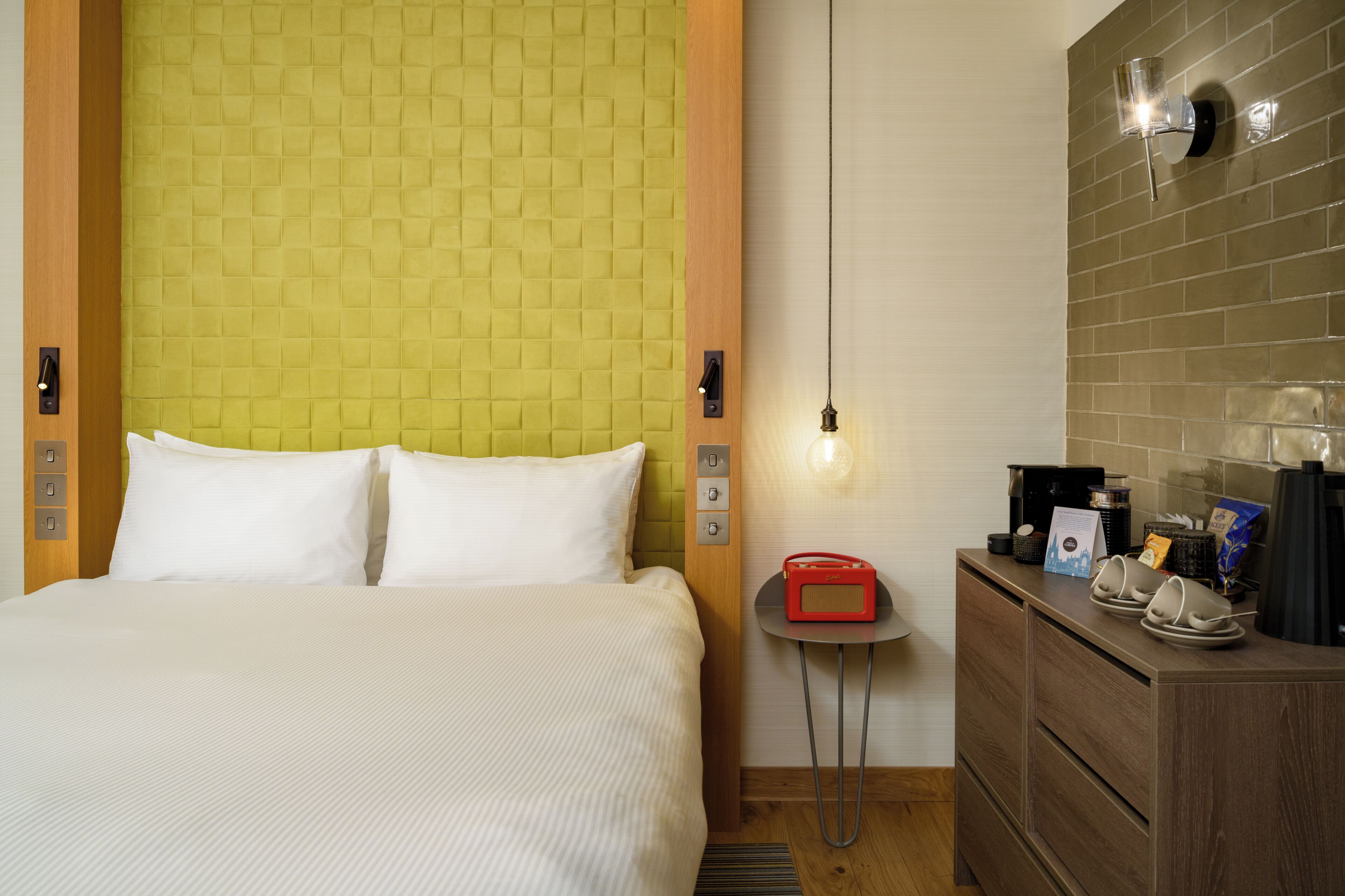 A hotel room with a bed, a small table with a radio, a coffee machine, and a wall-mounted light fixture.