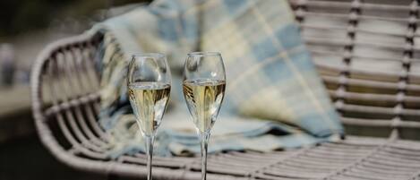 Deux verres de vin blanc sur une table, avec une chaise en osier et un coussin bleu et blanc en arrière-plan.