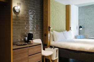 A hotel room with a bed, a desk with a coffee maker, a chair, and a nightstand.