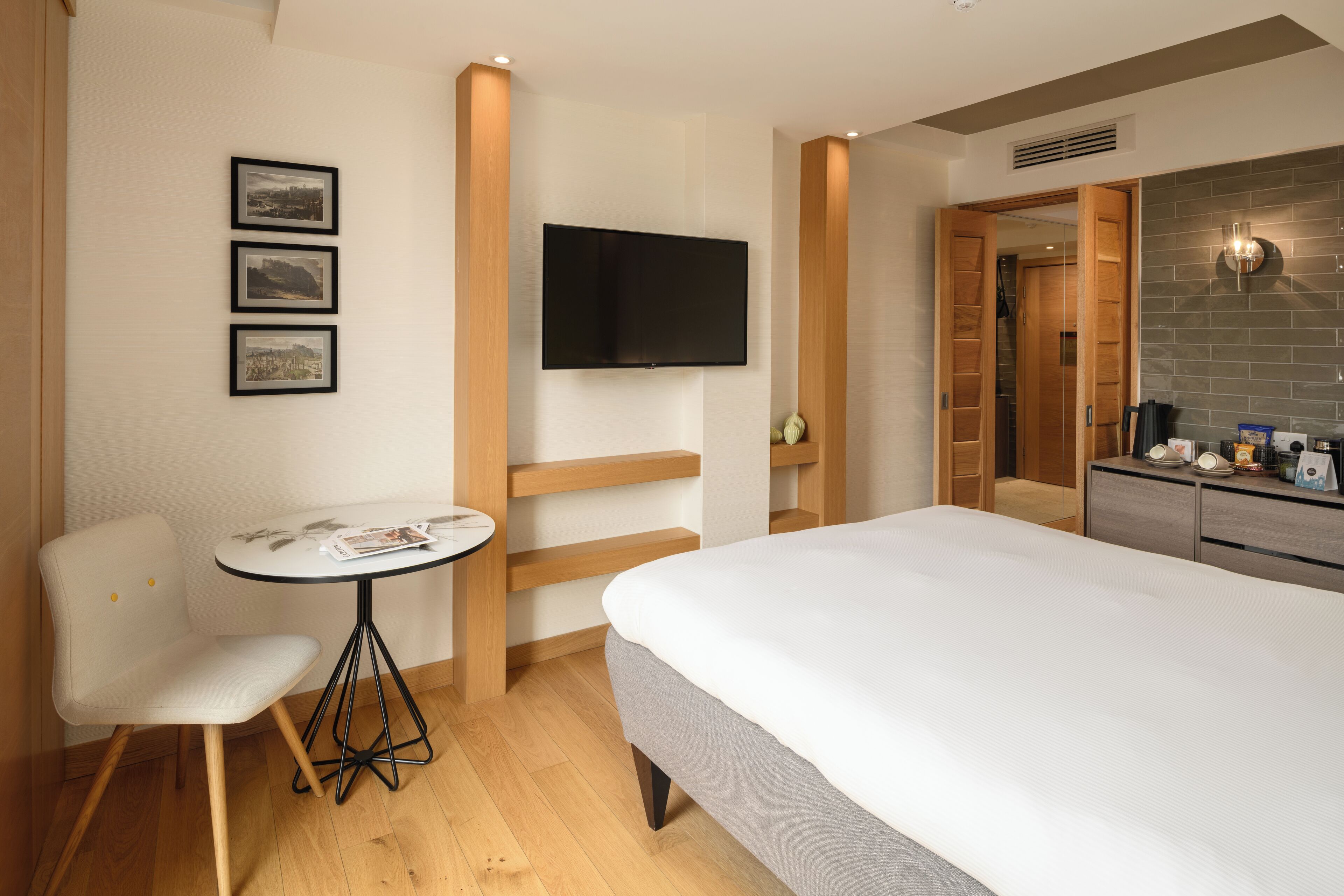 A hotel room with a bed, a small table with a chair, a TV mounted on the wall, and a kitchenette area.