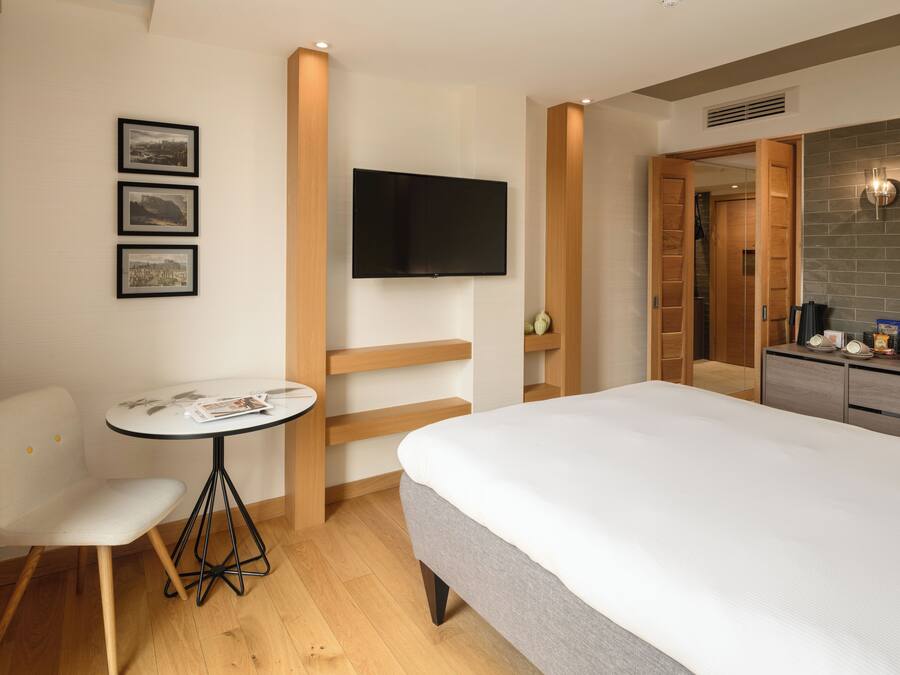 A hotel room with a bed, a small table with a chair, a TV mounted on the wall, and a kitchenette area.