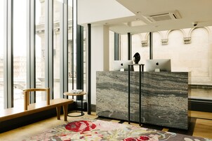 A modern hotel lobby with a marble reception desk, a wooden bench, and a view of the city through large windows.