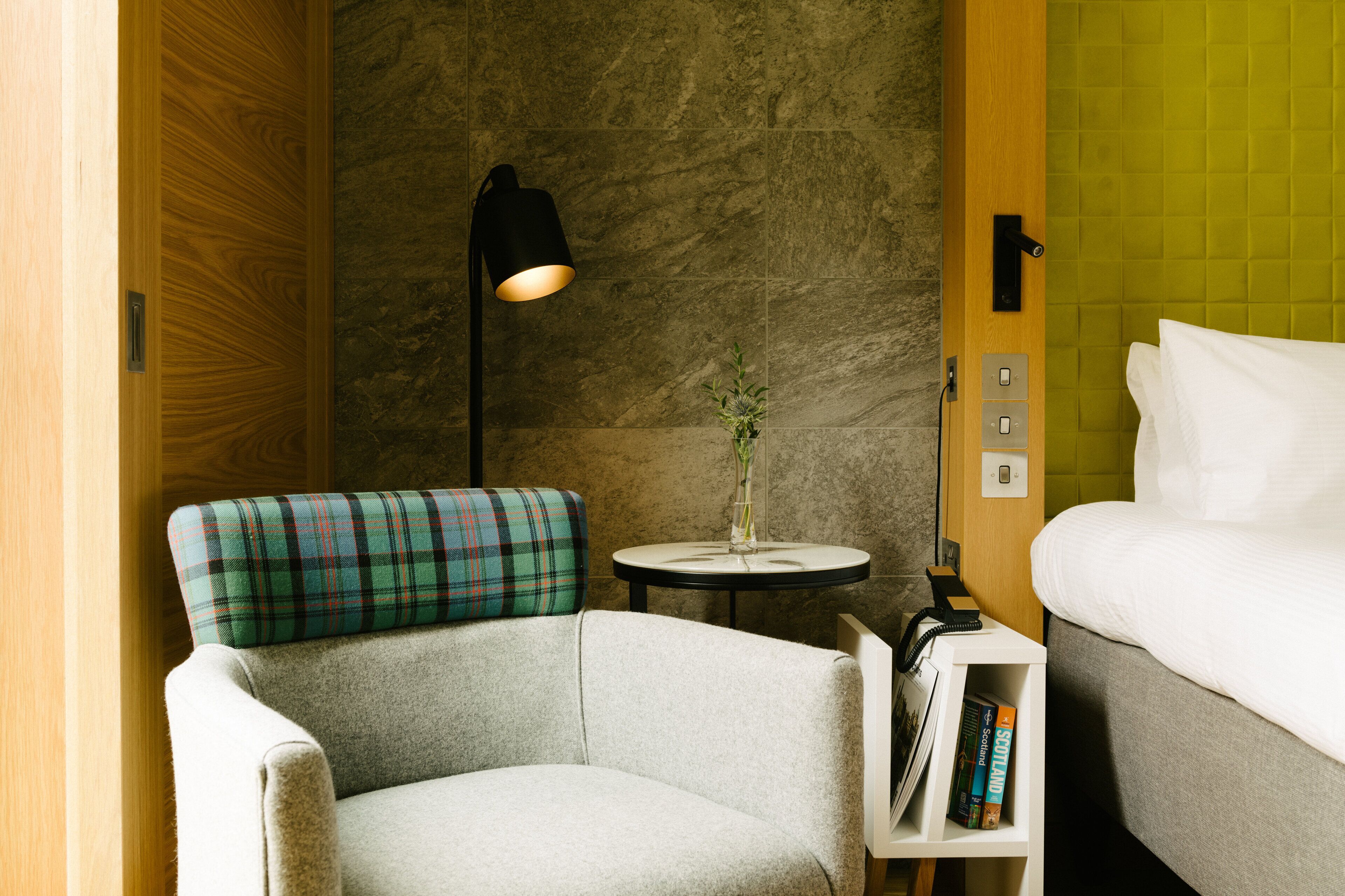 A hotel room with a plaid armchair, a bedside table with a lamp, a vase with a plant, and a bed with white linens.