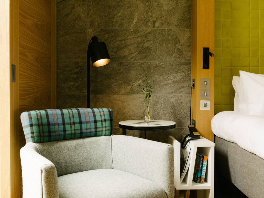 A hotel room with a plaid armchair, a bedside table with a lamp, a vase with a plant, and a bed with white linens.