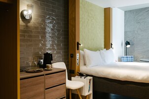 Een moderne hotelkamer met een bakstenen muur, een houten bureau met een koffiezetapparaat, een bed met wit beddengoed en een nachtkastje met een lamp.