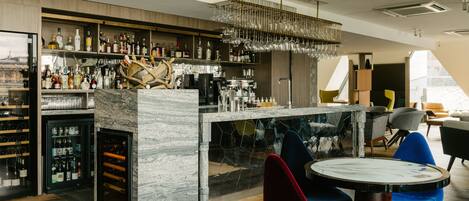 A modern bar area with a marble countertop, shelves of liquor bottles, and a wine cooler. There are colorful chairs and a round table in the foreground.