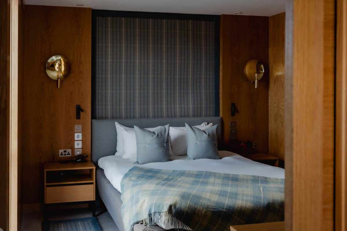 A bedroom with a bed, bedside tables, a nightstand, a wall-mounted lamp, and a window with blinds.