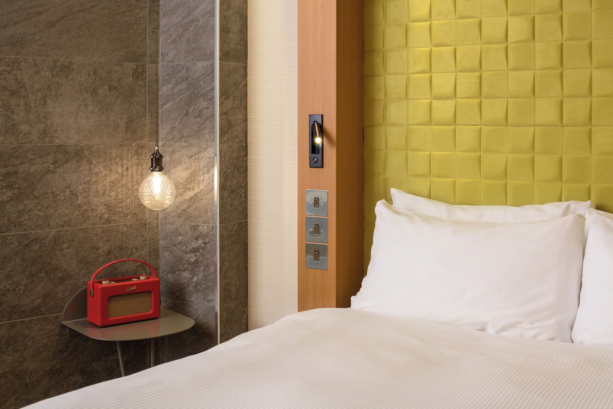 A hotel room with a bed, a bedside table with a red radio, a light fixture, and a wall with a textured panel.