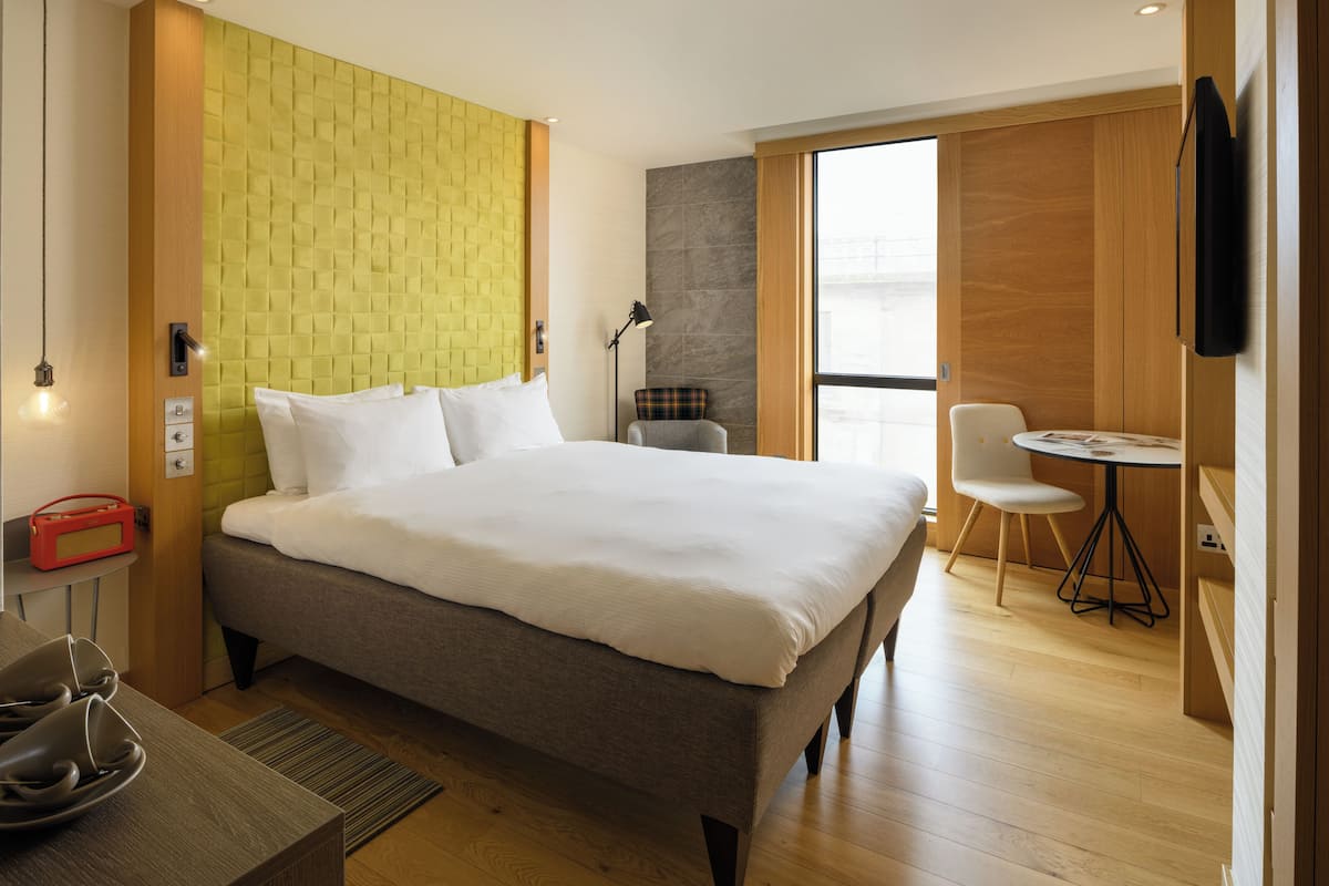 A modern hotel room with a large bed, a small table, a chair, and a TV mounted on the wall.