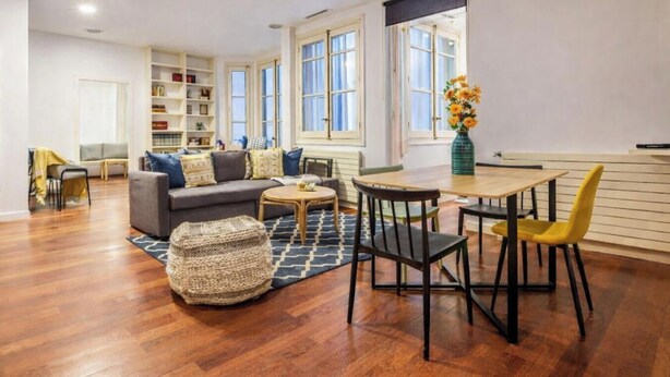 Una sala de estar con sofá, mesa de centro, mesa de comedor con sillas, estantería y ventana con vistas.