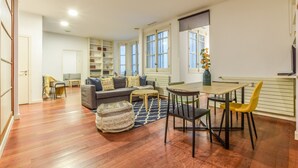 Amplia sala de estar con sofá, mesa de centro y mesa de comedor. Se observan estanterías y una ventana con vistas.