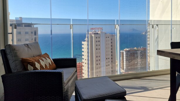 Een balkon met uitzicht op zee en gebouwen, een bank met kussens, en een kleine tafel.