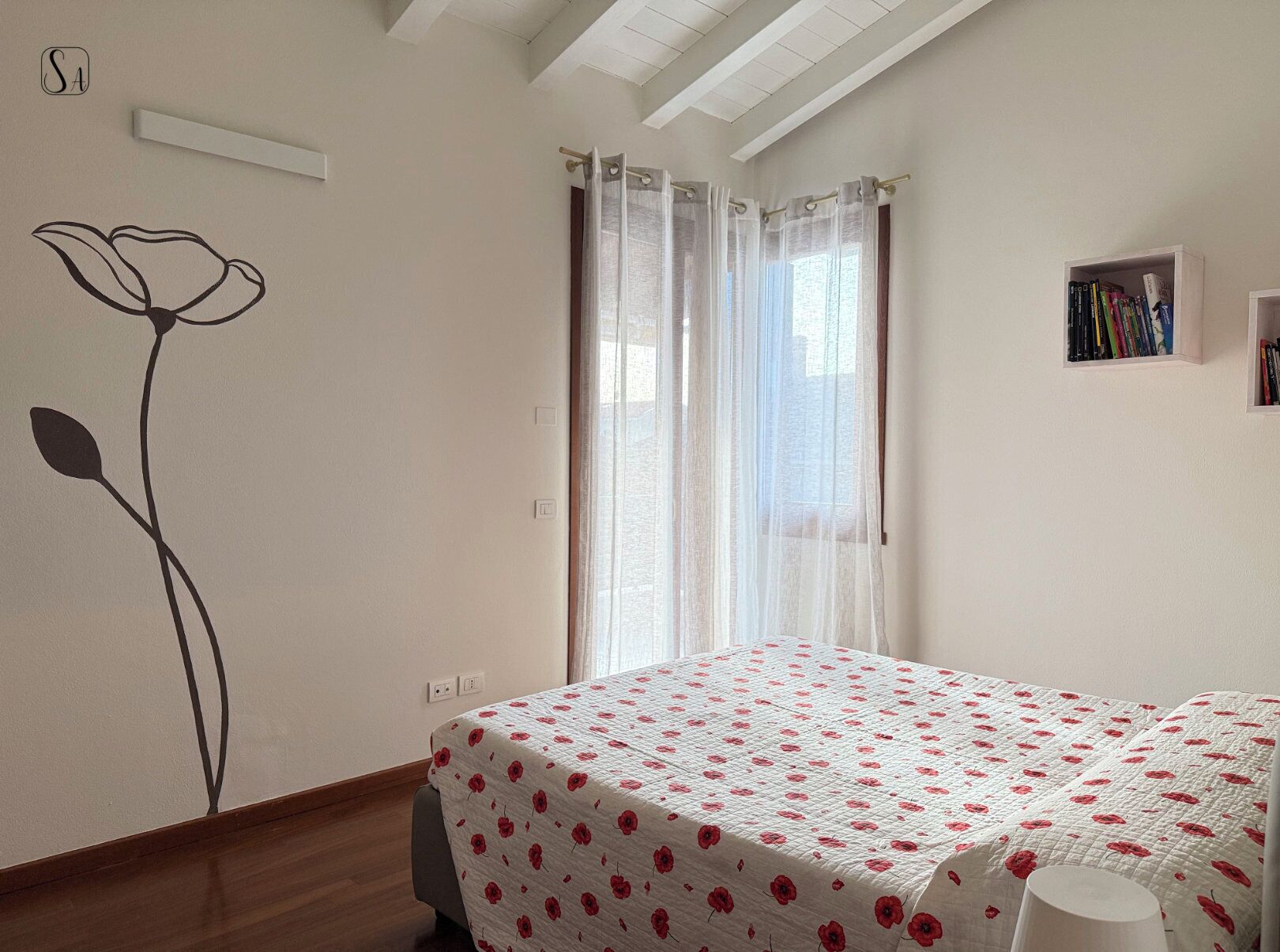 A bedroom with a bed, a window with sheer curtains, a wall with a floral design, and a small bookshelf.