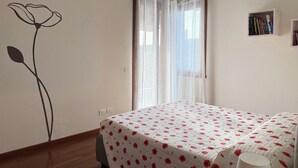 A bedroom with a bed, a window with sheer curtains, a wall with a floral design, and a small bookshelf. - Il Papavero apartment Sirmione | Terme & Garda Lake (Sirmione)