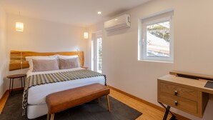 A modern bedroom with a wooden headboard, a bed with a patterned bedspread, a bench, a bedside table, a desk with drawers, and an air conditioning unit. - Very Nice Decorated 2 Bedroom Apartment (Oporto)