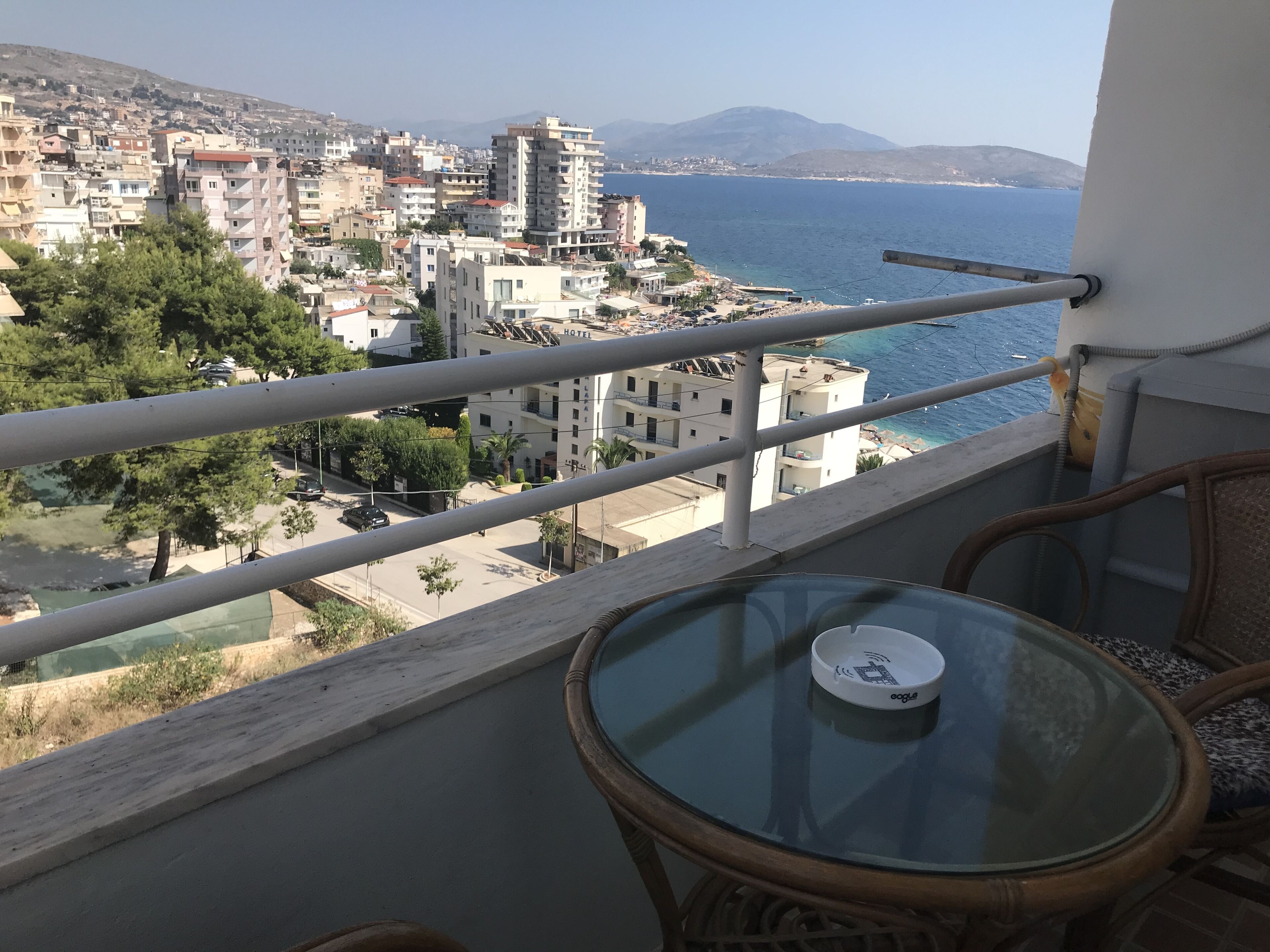 A balcony with a glass table and wicker chairs overlooking a coastal cityscape with buildings and a mountain in the distance.
