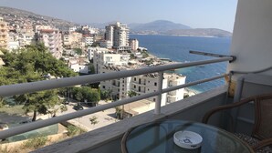 A balcony with a glass table and wicker chairs overlooking a coastal cityscape with buildings and a mountain in the distance. - Bella Apartment for rent near to the beach Sarandë , Albania  (Saranda)