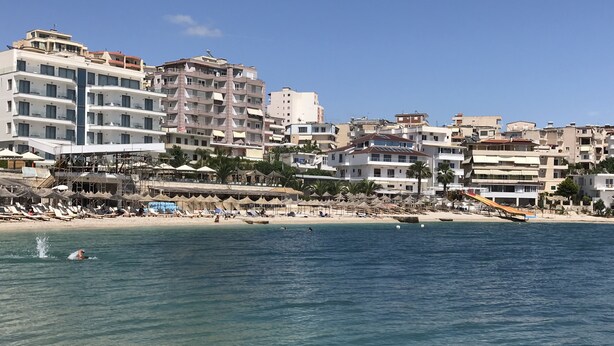 A coastal cityscape with a beach, including buildings, umbrellas, and a lifeguard tower. - Bella Apartment for rent near to the beach Sarandë , Albania  (Saranda)