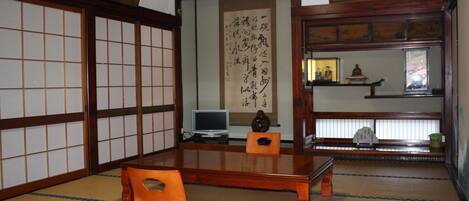 A traditional Japanese room with tatami flooring, a low wooden table, and a tatami mat chair. There is a television on the table, a framed calligraphy scroll on the wall, and a small shrine with a statue.