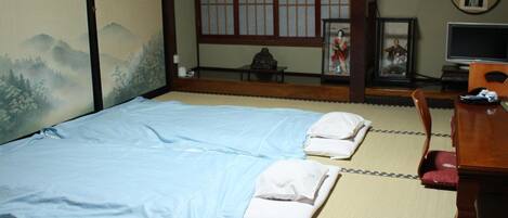 A traditional Japanese-style room with tatami flooring, a low wooden bed with blue bedding, a small table, and a TV.