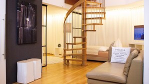 A modern living room with a wooden spiral staircase, a beige sofa, and a white pouf with text. - Best deal in town ! Como Designer Apt deluxe in the center of Munich (Munich)