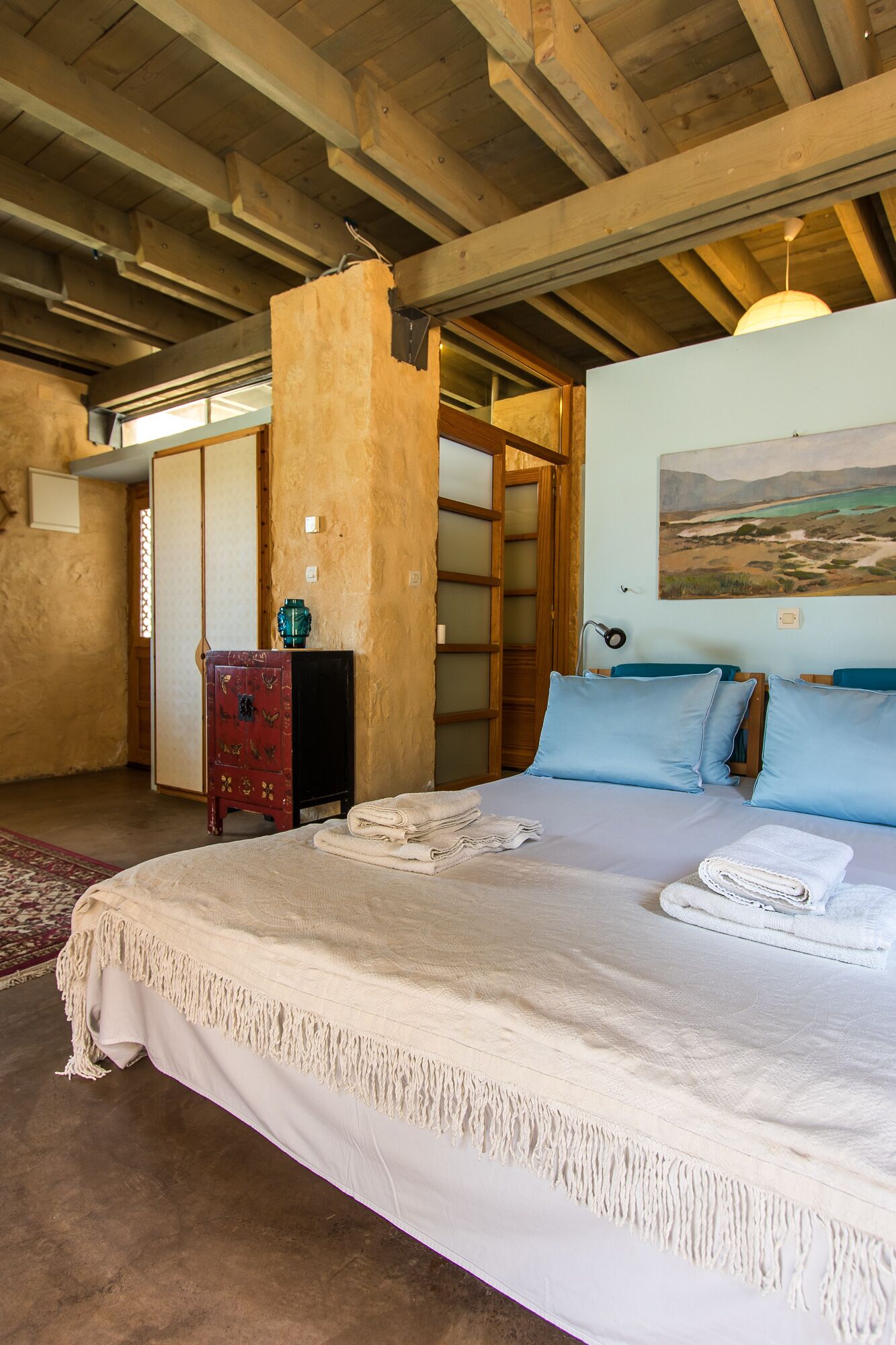 Een slaapkamer met een groot bed, een houten hoofdbord, een klein nachtkastje in rode tint, een schilderij aan de muur en uitzicht op het omliggende landschap via een deur.