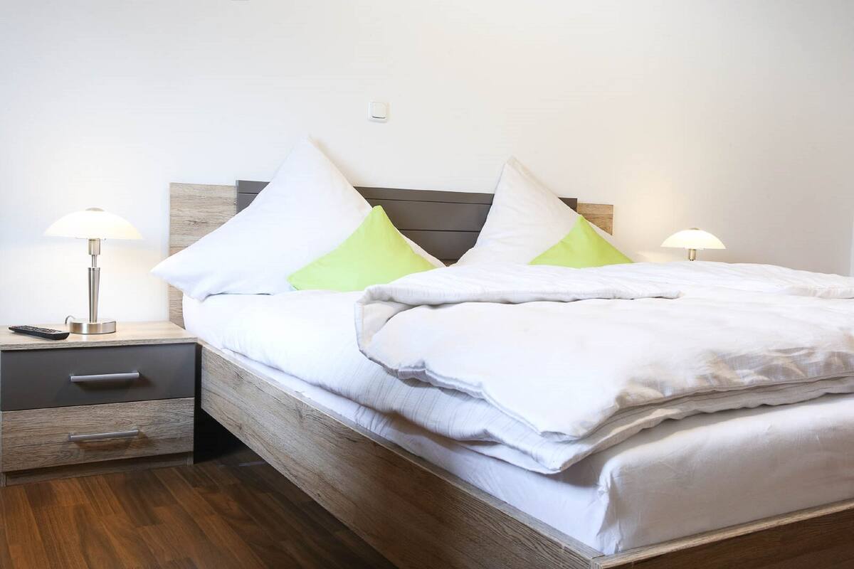 A neatly made bed with white and green pillows, a wooden headboard, and a bedside table with a lamp.