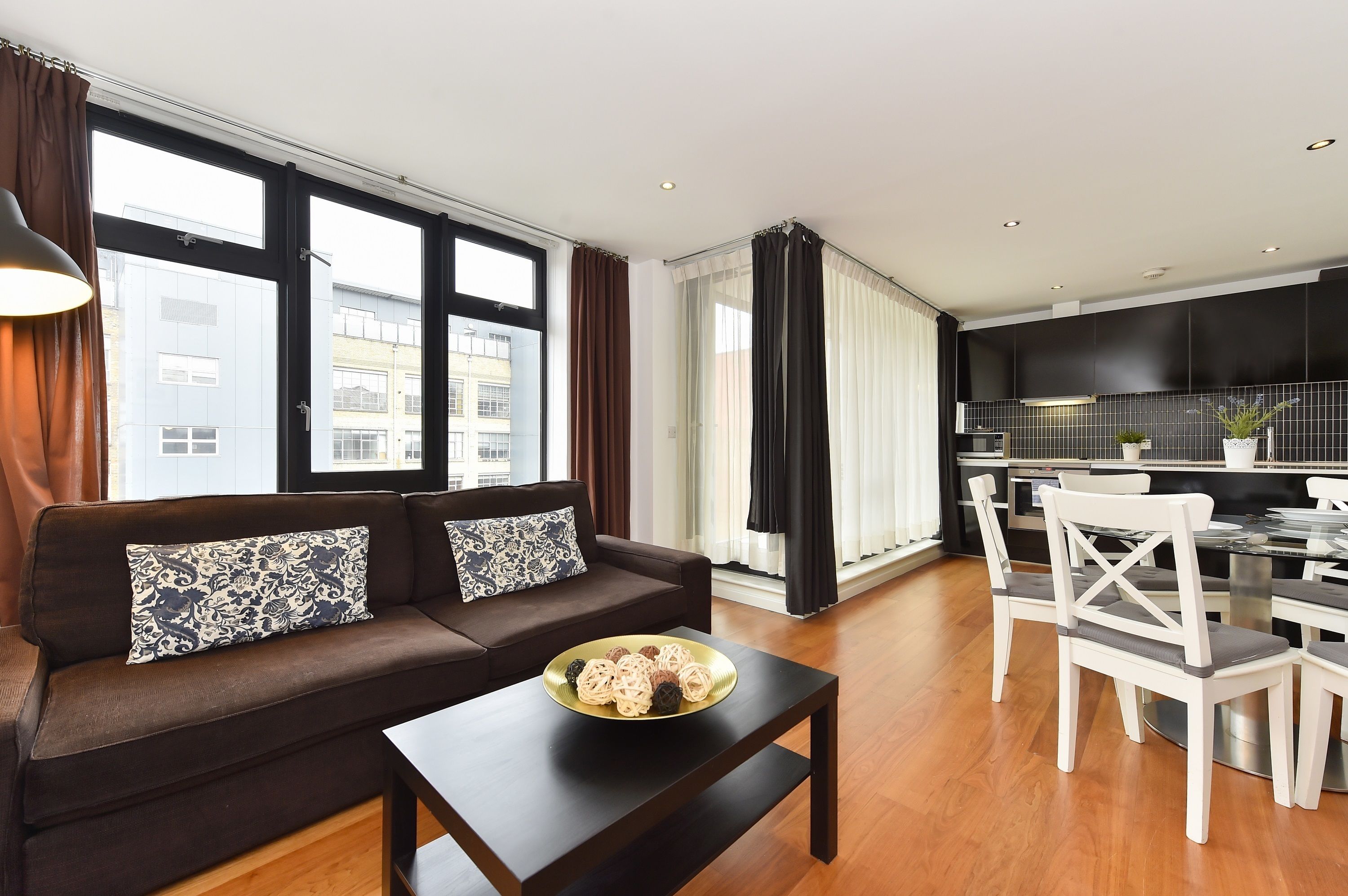 A modern living room with a sofa, a dining table, and chairs. There is a window with curtains, a kitchen area with cabinets, and a view of buildings outside.