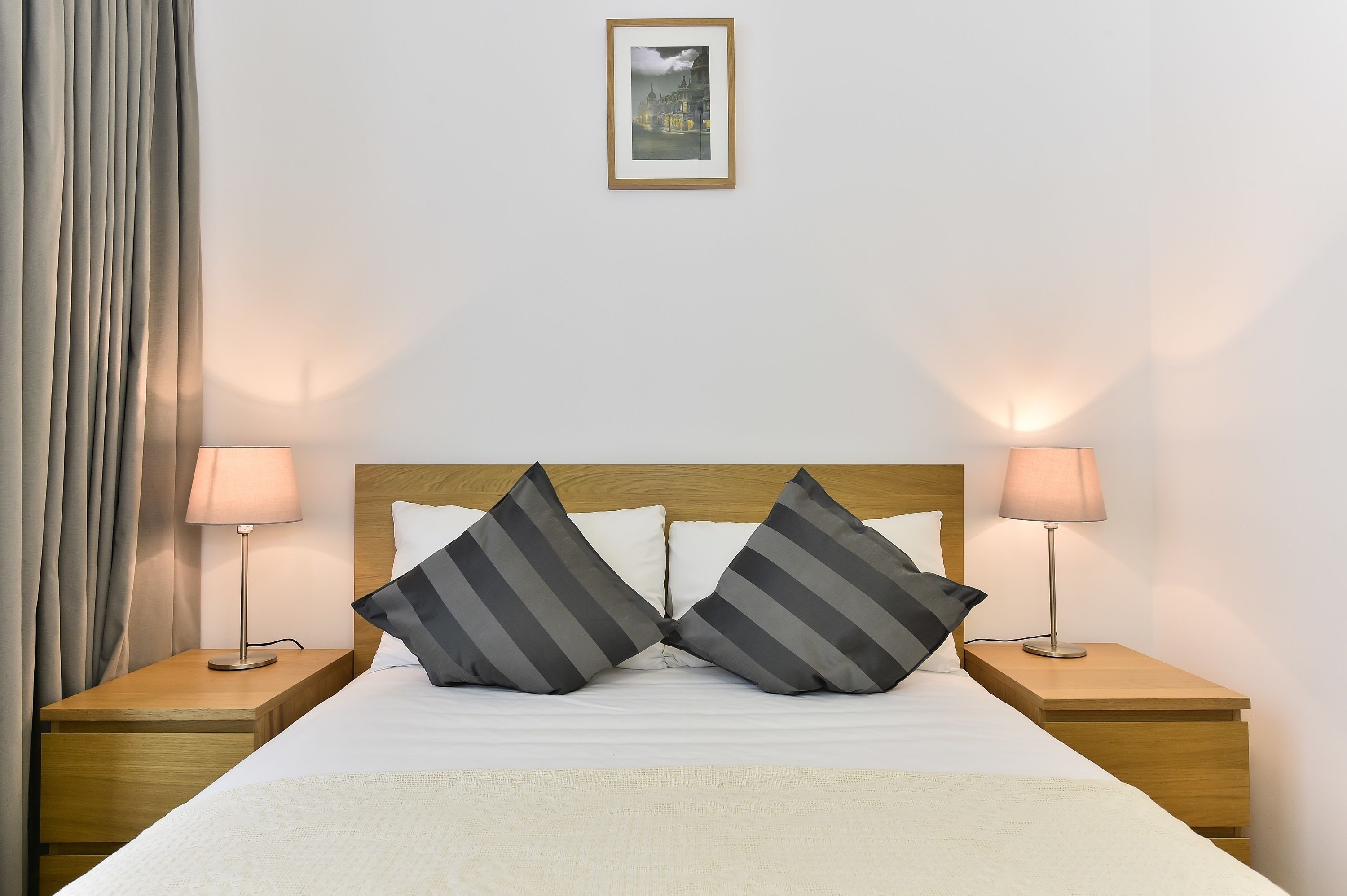 A neatly made bed with two pillows, two bedside lamps, a wooden headboard, and a framed picture on the wall.