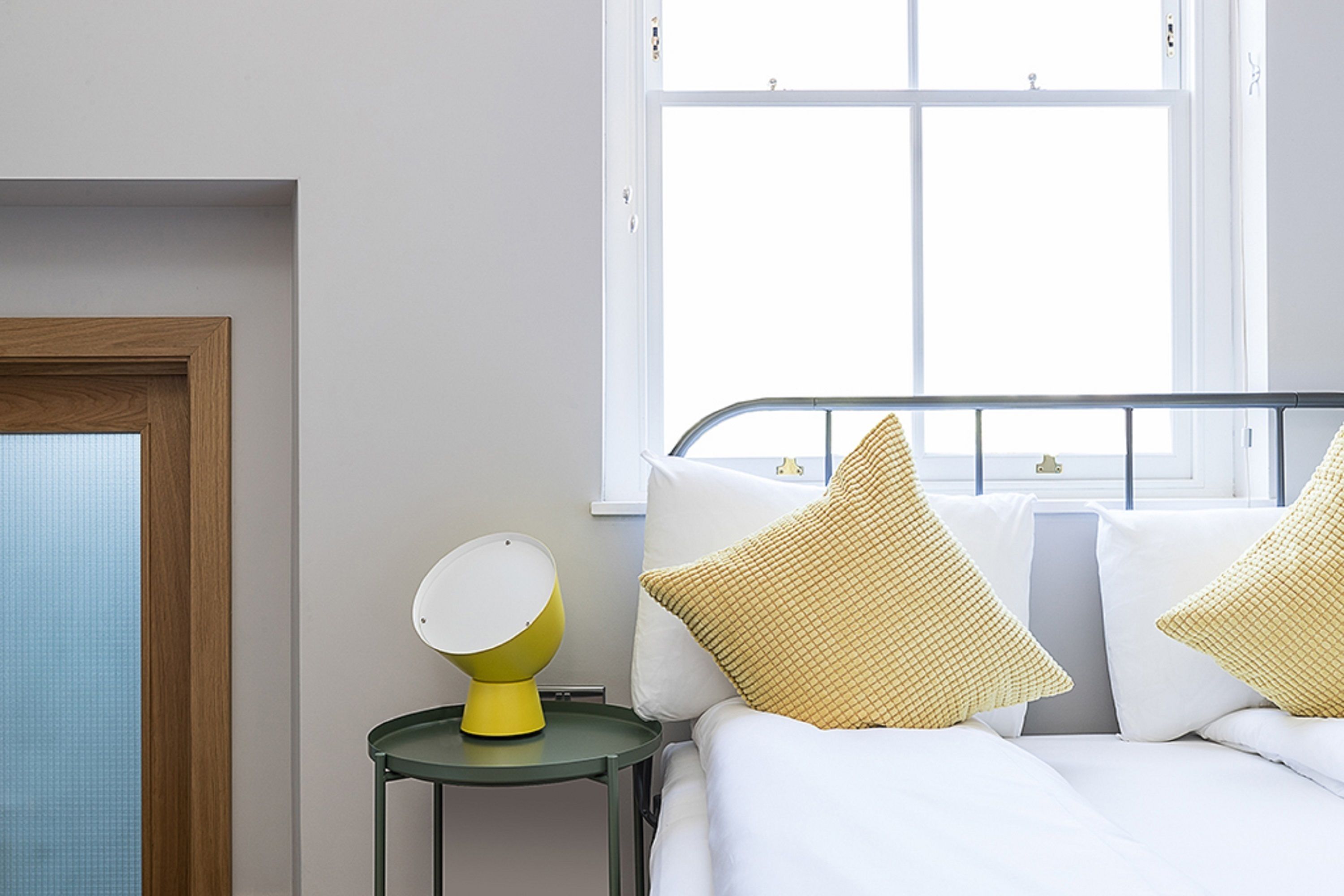 Een slaapkamer met een bed, kussens, een nachtkastje met een lamp en een raam.