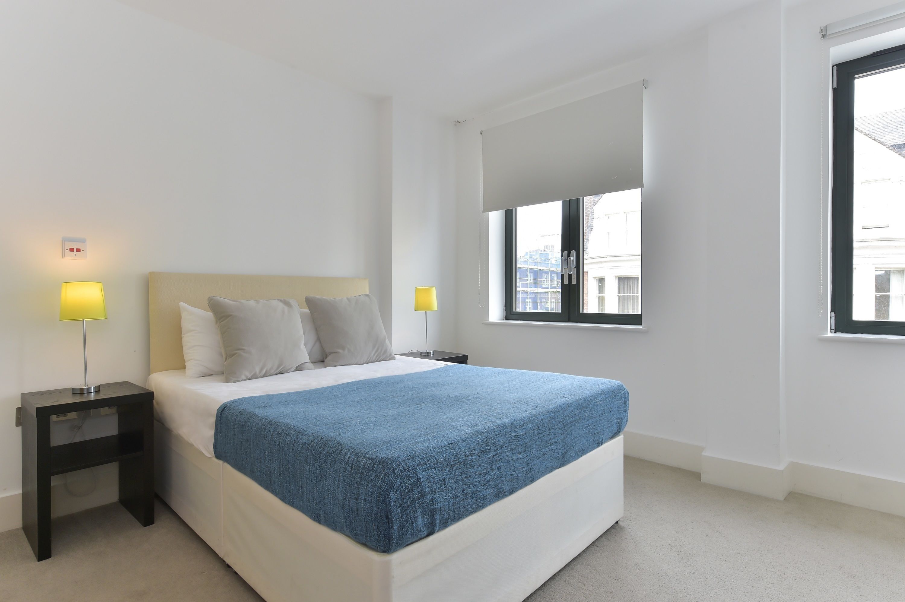Une chambre à coucher moderne, avec un lit, deux tables de chevet dotées de lampes, une grande fenêtre et une vue sur les bâtiments environnants.