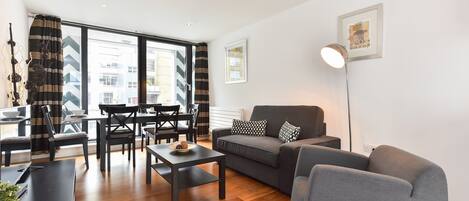 A modern living room with a grey sofa, black and white patterned curtains, a dining area with a table and chairs, and a large window offering a city view.