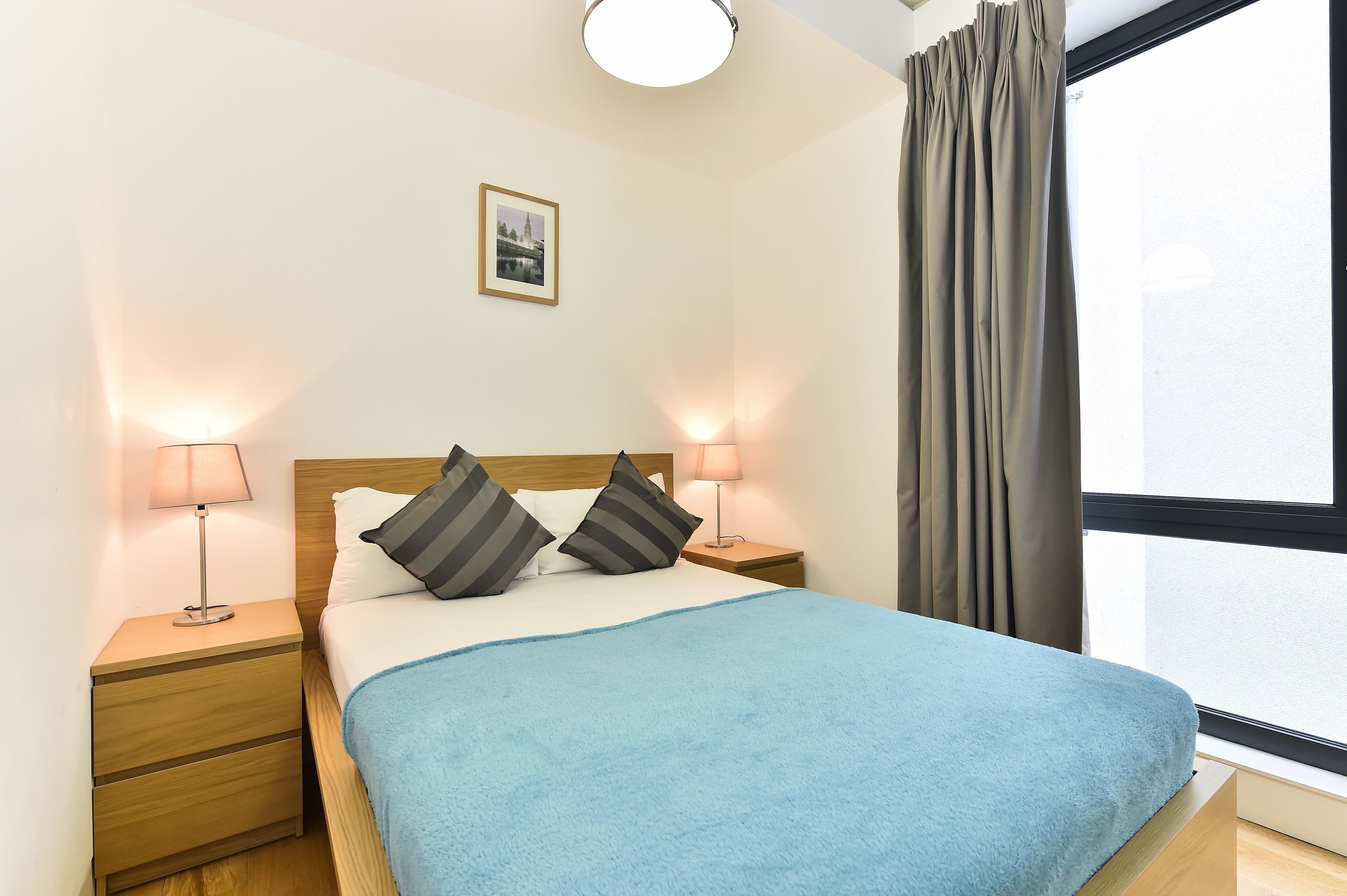 A neatly made bed with a blue blanket, two bedside lamps, and a framed picture on the wall.