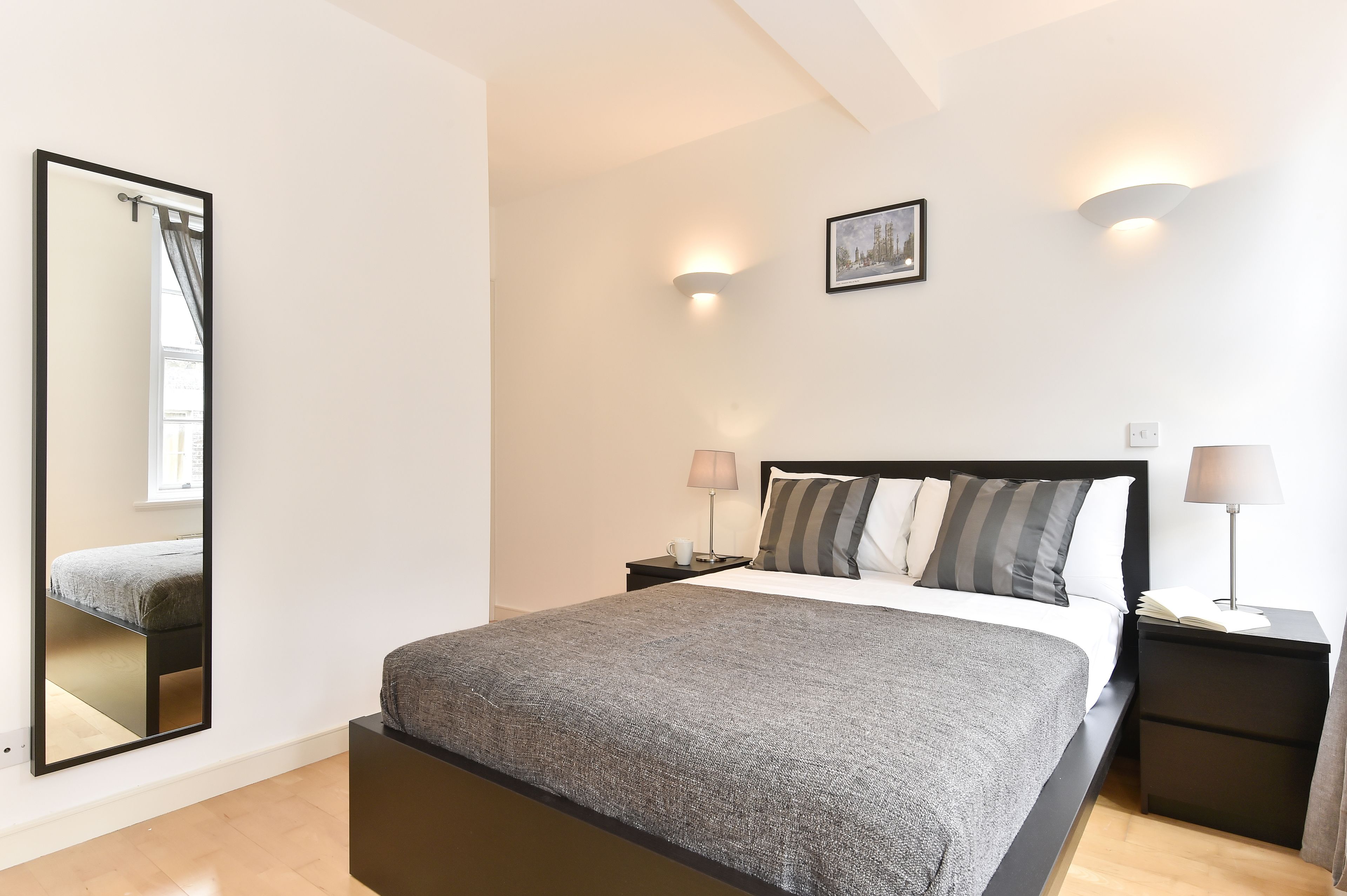 A bedroom with a large bed, a nightstand, a bedside lamp, a wall-mounted mirror, and a framed picture on the wall.