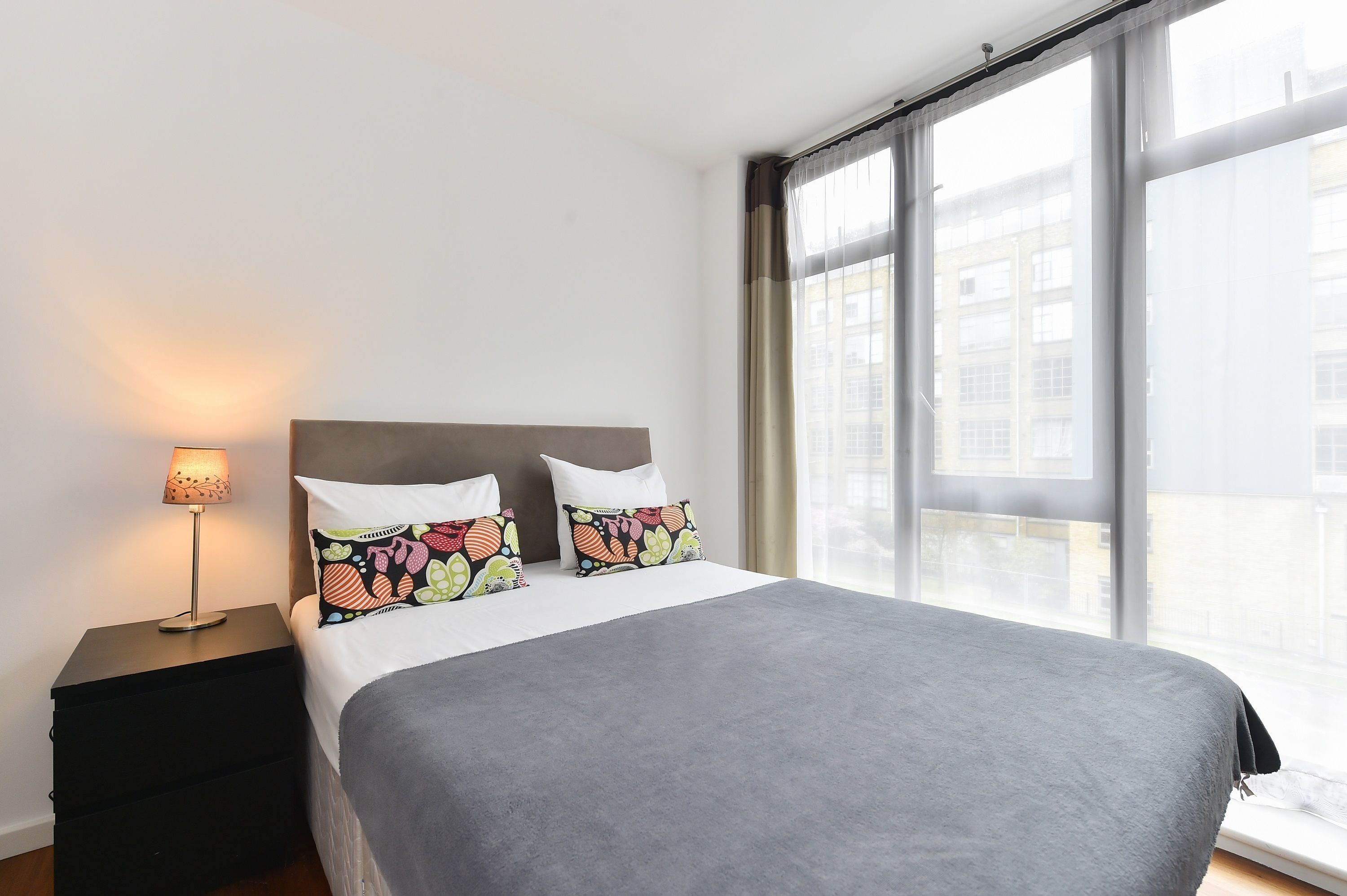 A hotel room with a large bed, two pillows with floral patterns, a bedside lamp, and a view of buildings through large windows.