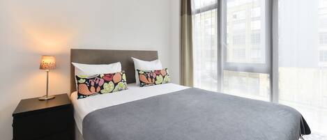A hotel room with a large bed, two pillows with floral patterns, a bedside lamp, and a view of buildings through large windows.