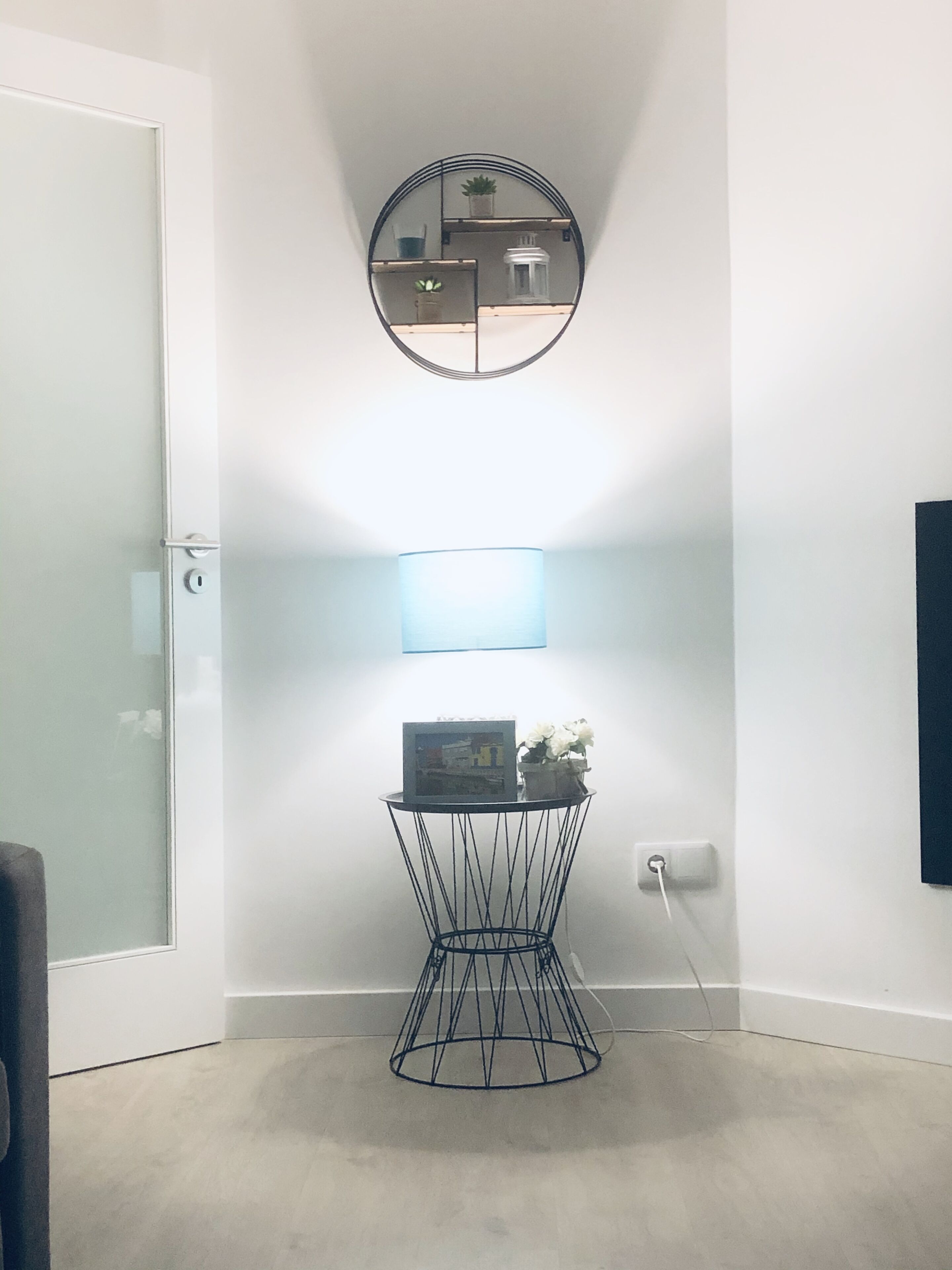 A modern room with a glass door, a round mirror, a small table with a picture frame and flowers, and a wall-mounted light.