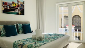 Ein Hotelzimmer mit einem Bett, einem Gemälde einer Haflandschaft, einem Balkon mit Blick auf ein Gebäude und einem Vorhangfenster.